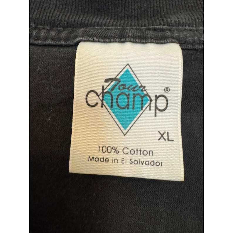 Puede incluir: Primer plano de una etiqueta de camiseta negra. La etiqueta presenta el texto "Tour Champ" en una fuente estilizada, con una forma de diamante turquesa. La etiqueta tambi&eacute;n dice "XL", "100% Algod&oacute;n" y "Made in El Salvador".