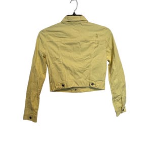 May include: A pale yellow denim jacket with long sleeves, a button-down front, and two chest pockets. The jacket is hanging on a black hanger against a white background. The jacket has a distressed look.