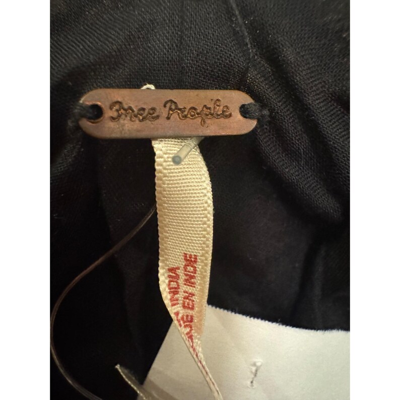 May include: A close-up of a black garment with a brown tag that reads "Free People." A cream-coloured tag with the text "INDIA" and "EN INDE" is attached. The garment appears to be made of a soft, dark fabric.