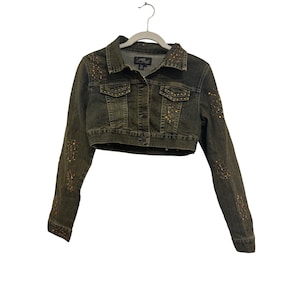 May include: Cropped olive green denim jacket with button closure, two chest pockets, and embellished with gold and bronze studs and rhinestones. The jacket is hanging on a white hanger.