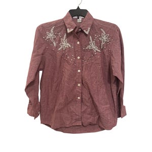 May include: A long-sleeved button-down shirt in a dusty rose color, featuring a pointed collar and a front button placket. The upper chest and collar are decorated with white beadwork and embroidery in floral and vine-like patterns.