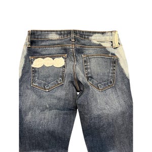 May include: Blue denim jeans with a light cloud-shaped patch on the back pocket. The jeans have a faded wash and a classic five-pocket design. The back of the jeans is visible, showing the pockets and waistband.