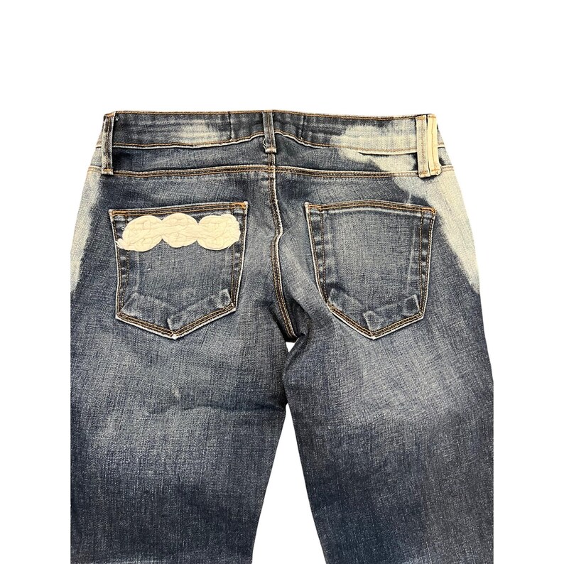 May include: Blue denim jeans with a light cloud-shaped patch on the back pocket. The jeans have a faded wash and a classic five-pocket design. The back of the jeans is visible, showing the pockets and waistband.