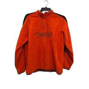 May include: Orange fleece Nautica Jeans pullover with a black zipper and black accents on the shoulders. The Nautica Jeans logo is printed in black across the chest. The garment is hanging on a black hanger.