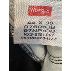 Op de afbeelding: Close-up van een wit label met het rode "Wrangler" logo. Het label toont de maat "44 X 30" en verschillende productcodes. Het label is genaaid op een donkere spijkerbroek.