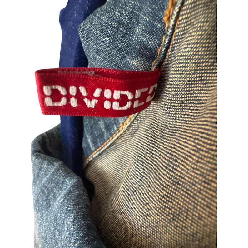 May include: A close-up of a blue denim jacket with a red fabric label that says "DIVIDED".