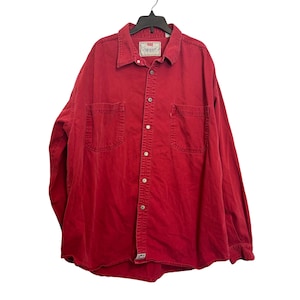 Puede incluir: Camisa roja de manga larga con dos bolsillos en el pecho y cuello, colgada de una percha negra. La etiqueta de la marca Levi's es visible en el pecho. Cierre de botones.