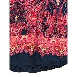 May include: Close-up of a fabric with a paisley pattern in shades of red, pink, and navy blue. The fabric features intricate floral and teardrop designs, with a decorative border at the bottom.