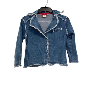Puede incluir: Chaqueta vaquera de lavado medio con cuello y bordes deshilachados. La chaqueta tiene una parte delantera abotonada, un pequeño bolsillo en el pecho y costuras decorativas en los hombros. La chaqueta está colgada de una percha negra.