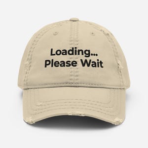 May include: A distressed beige baseball cap with the text "Loading... Please Wait" in black. The cap has a curved brim and a worn, vintage look, with intentional distressing around the edges.