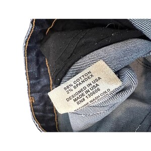 May include: Close-up of a denim garment with a white tag. The tag reads "98% Cotton, 2% Spandex, Designed in USA, Made in USA, RN# 130606, Machine Wash Cold."