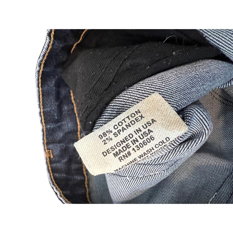 May include: Close-up of a denim garment with a white tag. The tag reads "98% Cotton, 2% Spandex, Designed in USA, Made in USA, RN# 130606, Machine Wash Cold."