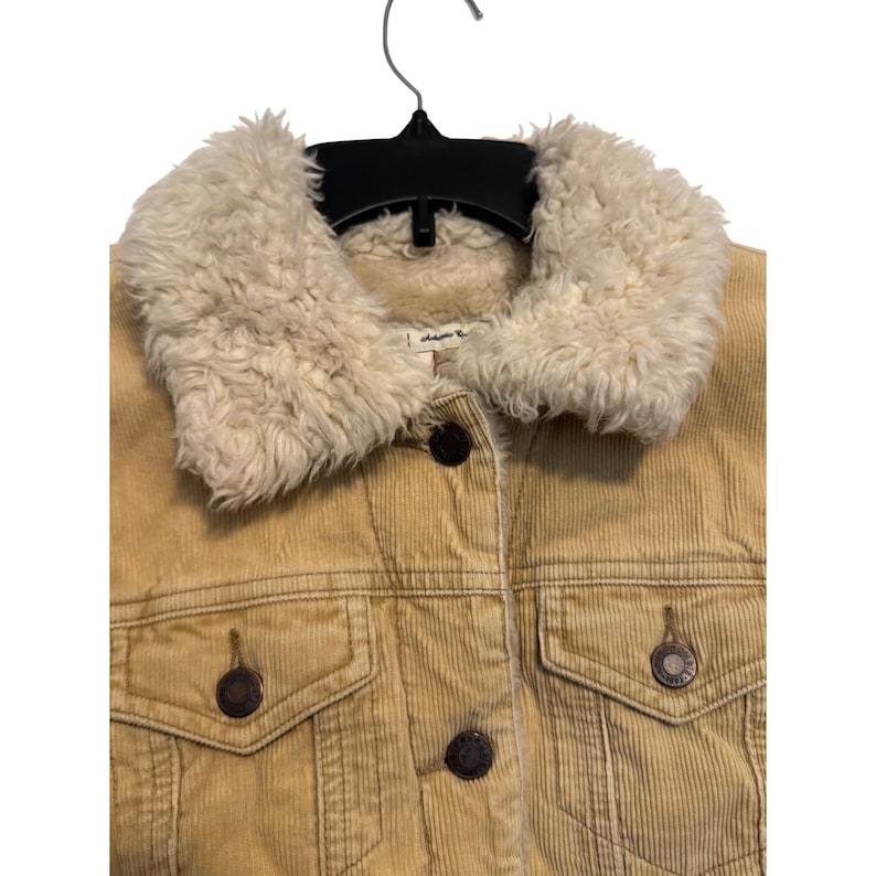 May include: A tan corduroy jacket with a cream-colored faux fur collar. The jacket has two front pockets, dark brown buttons, and is hanging on a black hanger. The jacket is a classic style.