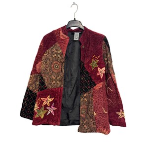 May include: A long-sleeve jacket featuring a patchwork design of deep red velvet, black fabric with cross-stitch patterns, and panels with intricate brown and gold paisley prints. The jacket includes embroidered leaf motifs and a black lining.