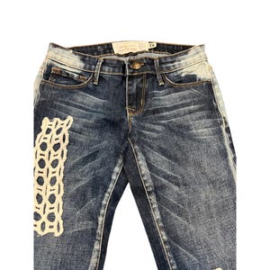 May include: Dark wash denim jeans with a distressed look. Features a cream-colored, open-weave patch on the left leg. The jeans have a button and zipper closure, with a small tag at the waist. Size 24.