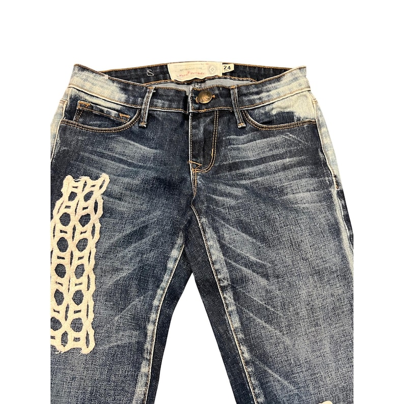 May include: Dark wash denim jeans with a distressed look. Features a cream-colored, open-weave patch on the left leg. The jeans have a button and zipper closure, with a small tag at the waist. Size 24.