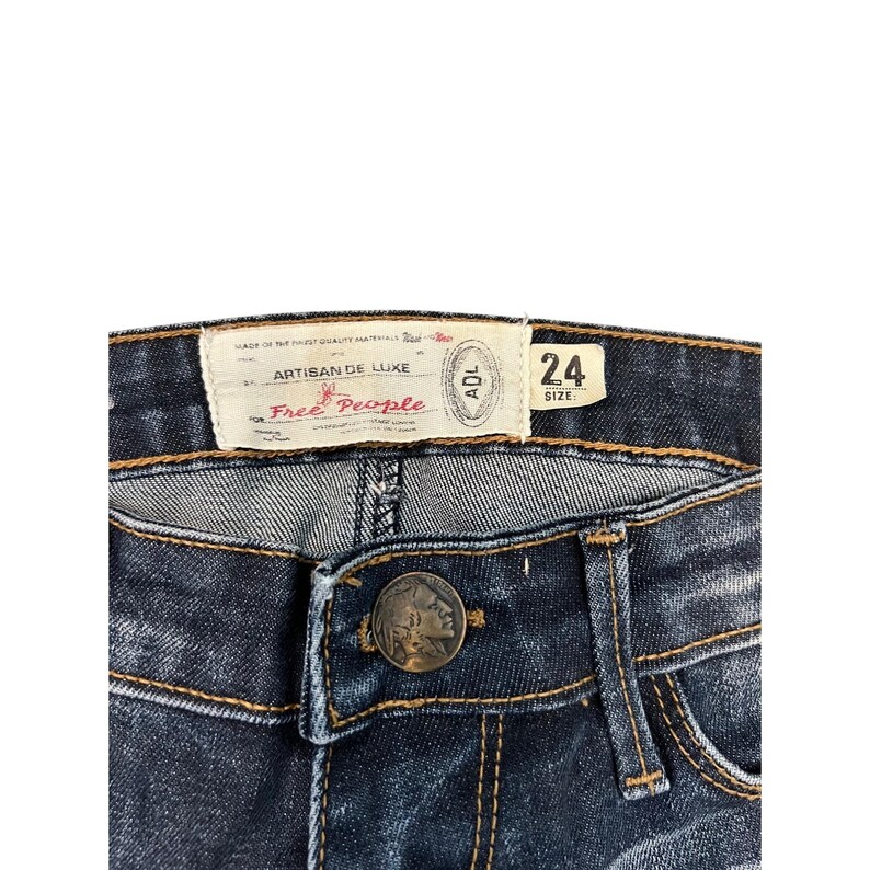 May include: Close-up of dark wash denim jeans. The jeans feature a button closure with a detailed coin-like button. A label reads "ARTISAN DE LUXE Free People" and the size is 24.