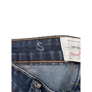 May include: Close-up of dark wash denim jeans. The jeans have a visible white "S" design and a white tag with the word "ARTIS" and the word "Free" in red.