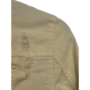 May include: A close-up view of a pale yellow denim jacket. The jacket has distressed detailing with ripped sections. Visible stitching and seams define the garment's construction. The fabric appears slightly worn.