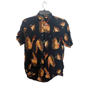 May include: Black short-sleeved shirt with a repeating pattern of brown, tan, and red feather-like shapes. The shirt is buttoned and hangs on a black hanger. The shirt has a single chest pocket.