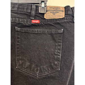Op de afbeelding: Close-up van een zwarte Wrangler-jeans. De jeans heeft een klassiek ontwerp met vijf zakken en een zichtbare achterzak. Een bruine leren patch met het Wrangler-logo staat op de achterkant. Het Wrangler-logo is ook zichtbaar op een rood label.
