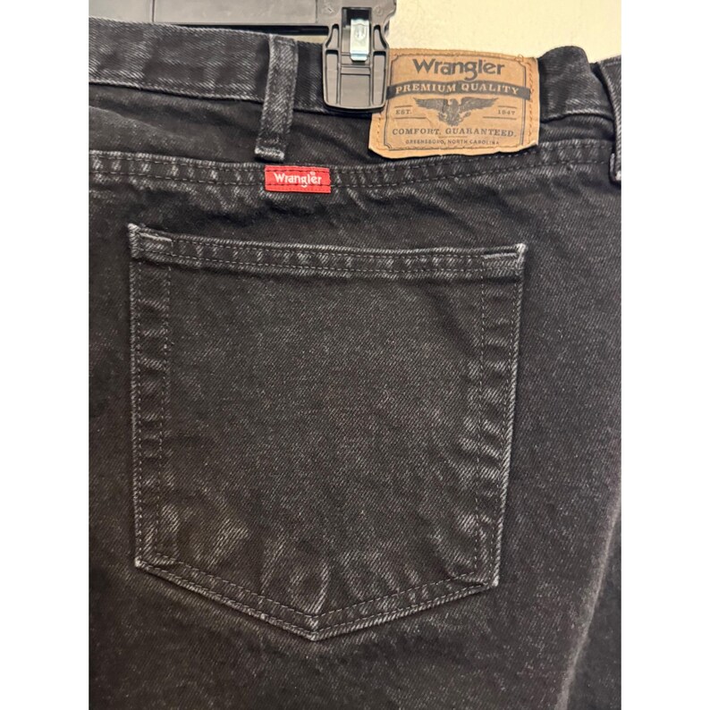 Op de afbeelding: Close-up van een zwarte Wrangler-jeans. De jeans heeft een klassiek ontwerp met vijf zakken en een zichtbare achterzak. Een bruine leren patch met het Wrangler-logo staat op de achterkant. Het Wrangler-logo is ook zichtbaar op een rood label.