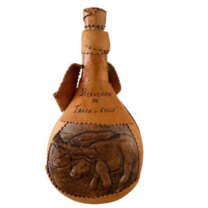 May include: A tan leather-covered bottle with a cork stopper, featuring a hand-tooled relief of two oxen pulling a cart. The text "Recuerdo de Talca - Chile" is inscribed on the upper section. The bottle has a narrow neck and a rounded base.