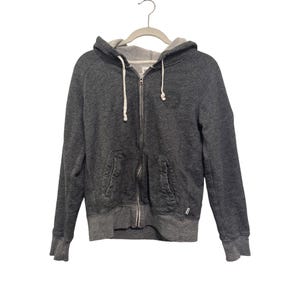 May include: A charcoal grey zip-up hoodie with a drawstring hood and two front pockets. The hoodie has a light grey interior and a small logo on the chest. It is hanging on a grey hanger.