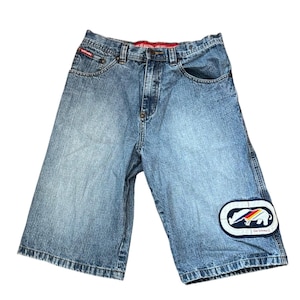 May include: Light blue denim shorts with a faded wash. Features a patch with a rhino logo and the text "Ecko Unltd." on the right leg. The shorts have a red waistband and classic five-pocket styling.