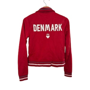 May include: Red zippered jacket with long sleeves and a white embroidered heart with a crown on the back. The text "DENMARK" is embroidered in white on the back of the jacket.