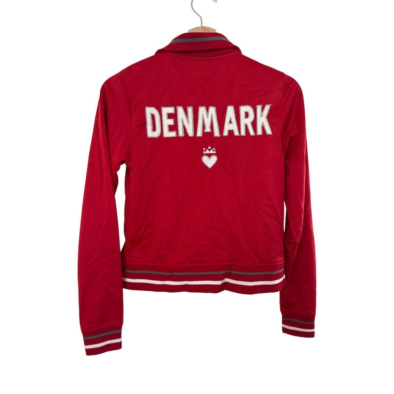 May include: Red zippered jacket with long sleeves and a white embroidered heart with a crown on the back. The text "DENMARK" is embroidered in white on the back of the jacket.