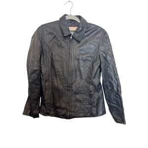 Puede incluir: Chaqueta de cuero marrón oscuro con cuello y cierre de cremallera completo. La chaqueta tiene un diseño clásico con un corte entallado y está colgada de una percha de metal. El cuero parece ligeramente desgastado, lo que aumenta su atractivo vintage.