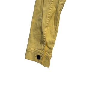 May include: Close-up of a yellow denim jacket sleeve. The sleeve has a buttoned cuff and a black button. The stitching is visible along the seams. The jacket is made of a soft, light-weight material.