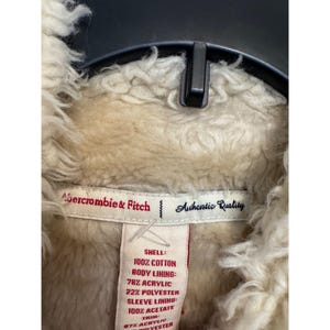 May include: Close-up of an Abercrombie & Fitch jacket tag. The tag reads "Authentic Quality" and lists the shell, body lining, sleeve lining, and trim materials. The jacket is cream-colored with a fuzzy texture.