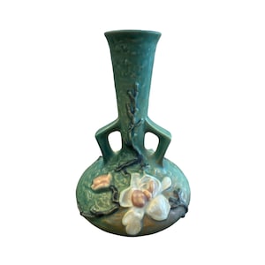 Op de afbeelding: Een blauwgroene keramieken vaas met een bolvormige voet en een uitlopende hals, voorzien van twee geïntegreerde handvatten. Het oppervlak is versierd met reliëfdetails van witte en roze bloemen en donkerbruine takken op een groene ondergrond.