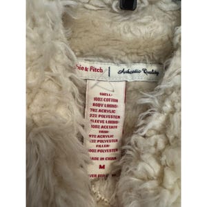 May include: Close-up of a cream-colored, textured jacket interior with a tag. The tag reads "Authentic Quality" and lists the materials: cotton, acrylic, polyester, and acetate. The jacket is a size M.