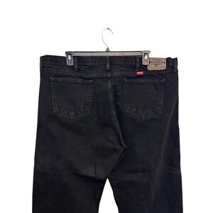Op de afbeelding: Zwarte Wrangler-jeans met een klassiek ontwerp met vijf zakken. De jeans hangen aan een zwarte hanger. Het Wrangler-merklabel is zichtbaar op de achterzak.