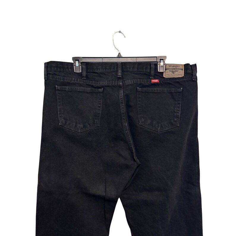 Op de afbeelding: Zwarte Wrangler-jeans met een klassiek ontwerp met vijf zakken. De jeans hangen aan een zwarte hanger. Het Wrangler-merklabel is zichtbaar op de achterzak.