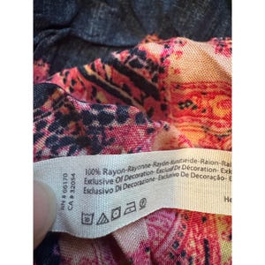 May include: Close-up of a patterned fabric with a tag. The fabric features a mix of pink, black, and orange hues. The tag displays text including "100% Rayon" and care symbols. The fabric is likely part of a garment.