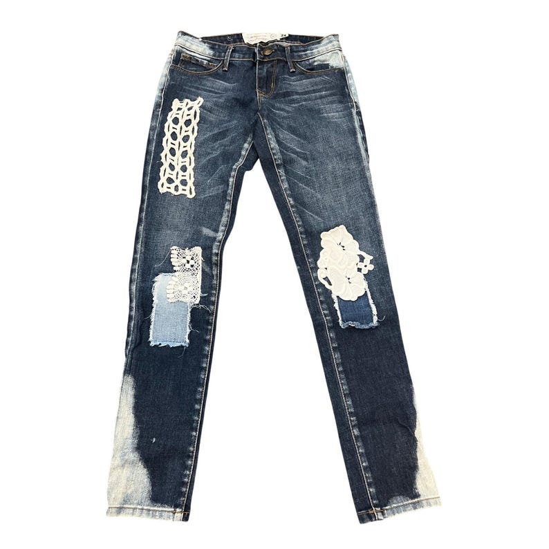 May include: Dark wash denim jeans with decorative white lace patches and distressed detailing. The jeans have a classic five-pocket design and a button and zipper closure. The bottom hems are faded.