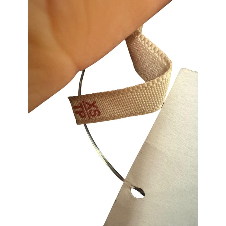 May include: Close-up of a clothing tag with a beige fabric loop and a white paper tag. The beige fabric loop has the letters "XS" printed in red. The paper tag is white and has a small hole.