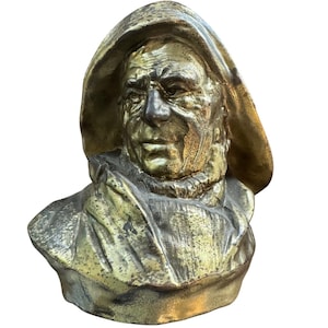 Puede incluir: Busto de bronce de un hombre con una prenda con capucha. La escultura tiene un aspecto envejecido y desgastado, con detalles visibles en el rostro y la ropa. El color es una mezcla de oro y bronce.