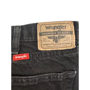 Op de afbeelding: Close-up van een zwarte denim jeans met een rood Wrangler-label en een bruine leren patch. De patch leest "Wrangler Premium Quality" met een adelaarslogo en "Comfort Guaranteed."