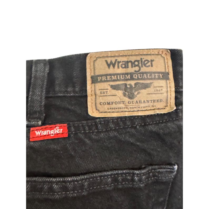 Op de afbeelding: Close-up van een zwarte denim jeans met een rood Wrangler-label en een bruine leren patch. De patch leest "Wrangler Premium Quality" met een adelaarslogo en "Comfort Guaranteed."