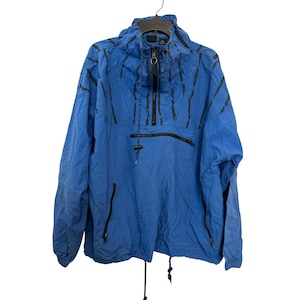 Puede incluir: Chaqueta anorak azul con capucha y cierre de cremallera. La chaqueta presenta detalles en negro, incluyendo rayas, cremalleras y cordones. Tiene dos bolsillos delanteros y está colgada de una percha.