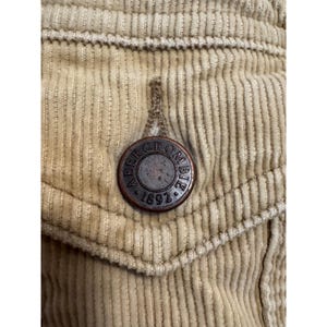 May include: Close-up of a tan corduroy fabric with a pocket and a metal button. The button is circular with the words "Abercrombie" and "1892" around the edge.