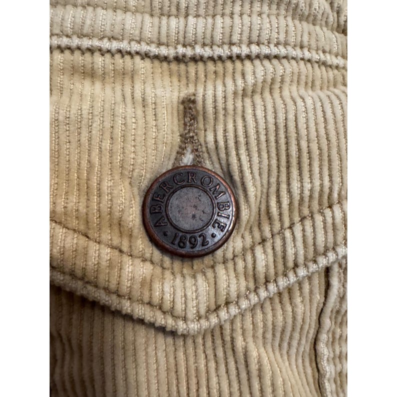 May include: Close-up of a tan corduroy fabric with a pocket and a metal button. The button is circular with the words "Abercrombie" and "1892" around the edge.