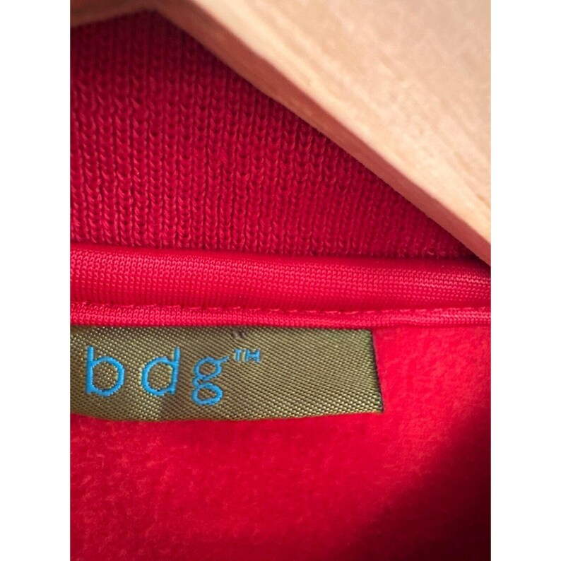 May include: Close-up of a red fleece jacket with a green label that says "bdg" in blue lettering.