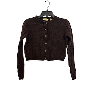 May include: A dark brown cable knit cardigan sweater with a round neckline and button-up front. The long-sleeved sweater is hanging on a black hanger against a white background. The sweater has a tag on the neckline.