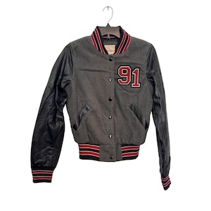 May include: Gray varsity jacket with black leather sleeves. Features a red and white striped collar and cuffs, and a large "91" patch on the chest. The jacket has a button-up front and two front pockets.